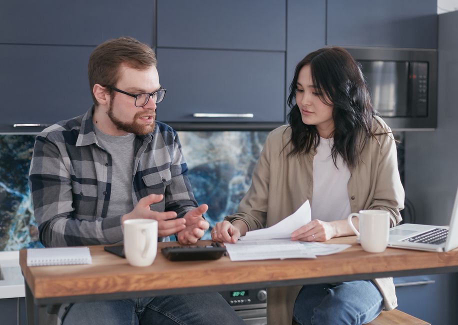 A couple sits at a bright, modern kitchen table with a laptop and calculator, thoughtfully reviewing documents and discussing the financial implications of selling their home.